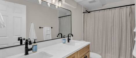 Une salle de bain moderne avec un grand miroir, deux vasques, des toilettes et un rideau de douche.