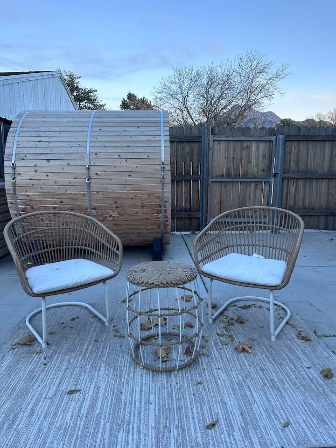 A patio area with two wicker chairs and a small round table, a wooden barrel, and a wooden fence in the background.