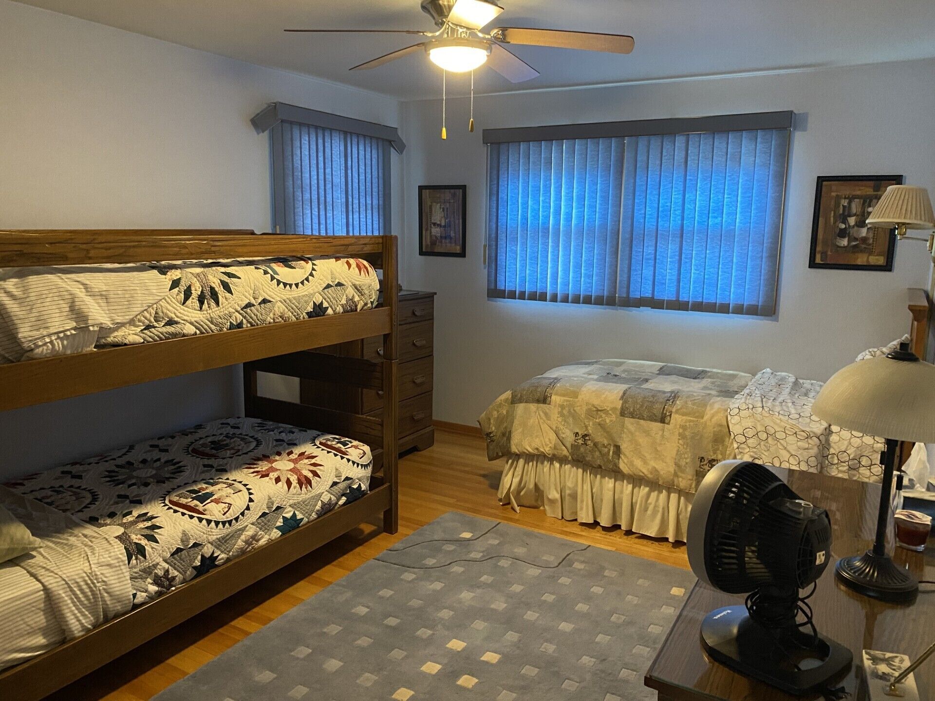 Una habitación con literas, un escritorio con lámpara, un ventilador y una ventana con cortinas.