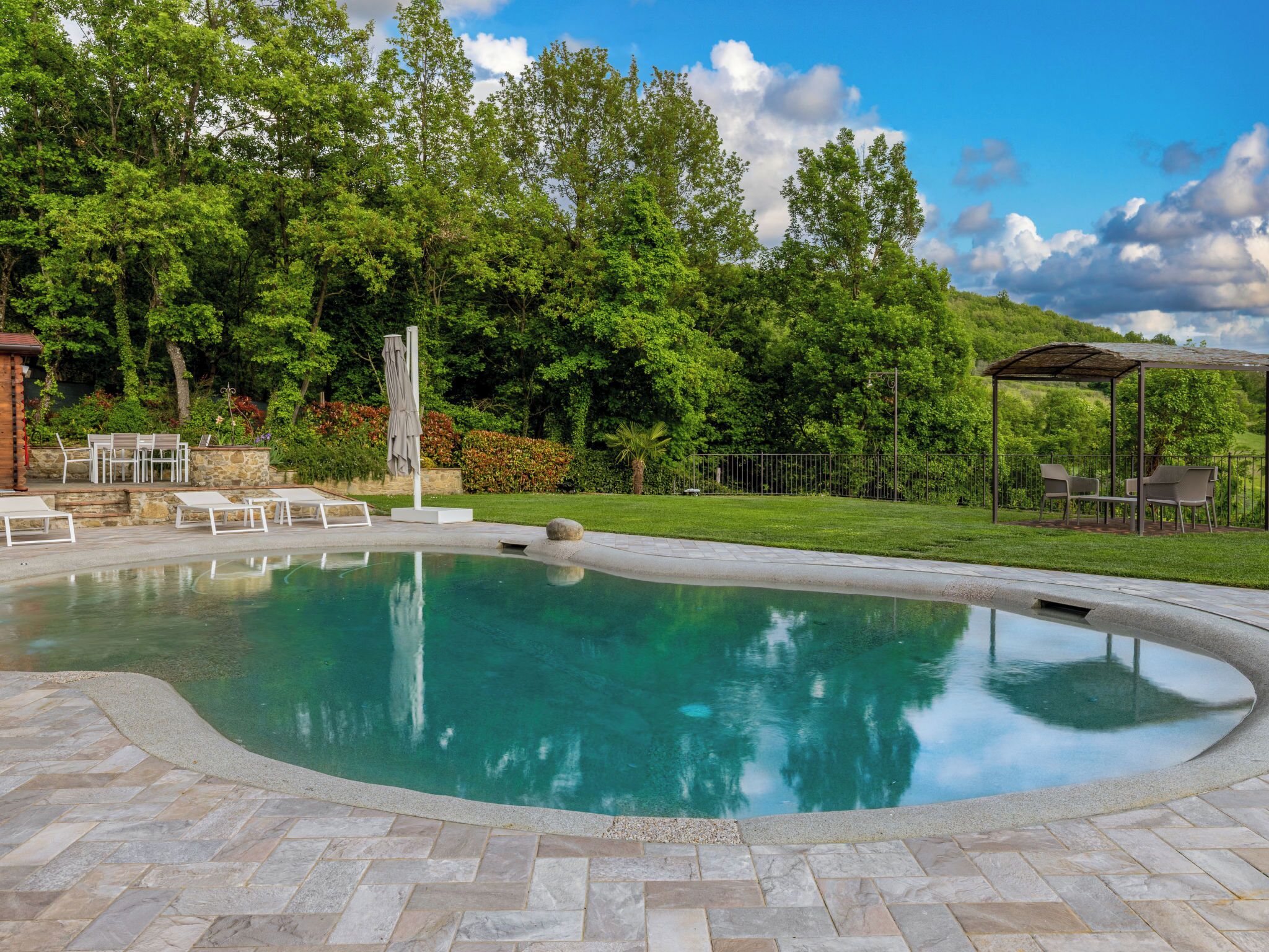 Una piscina con terraza de piedra, rodeada de follaje y un pabellón.