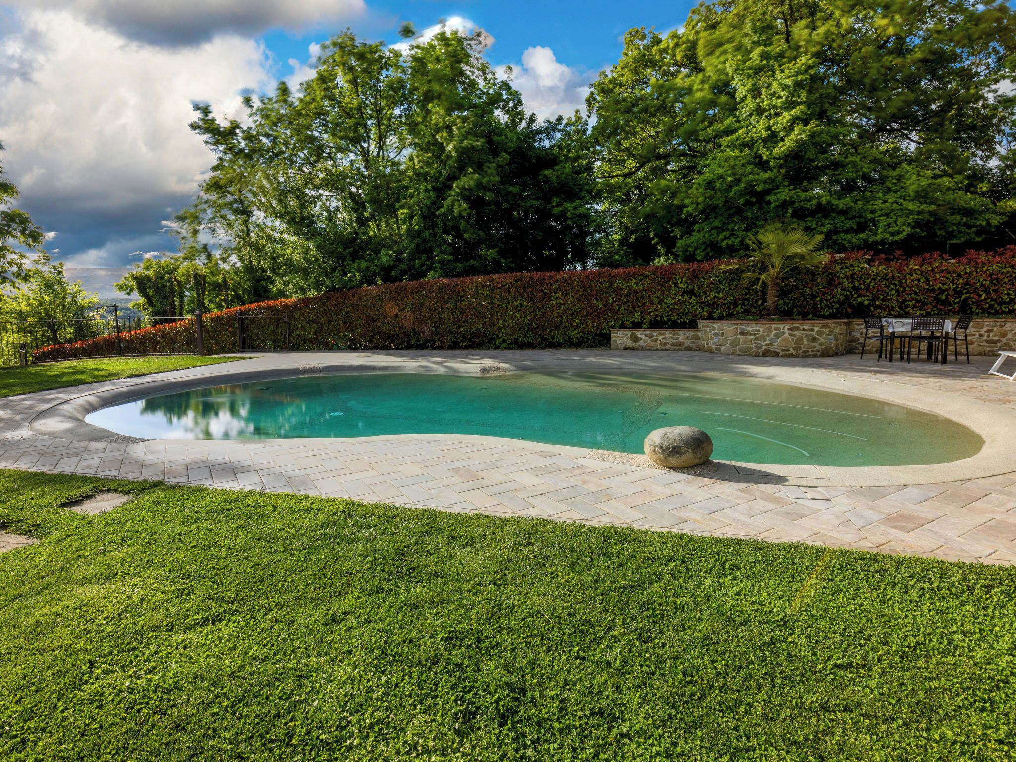 A swimming pool with a curved edge, surrounded by a stone wall and a hedge.