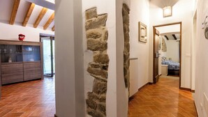 Un pasillo con pared de piedra, vigas de madera, una zona de cocina con armarios y una cama en la habitación contigua.