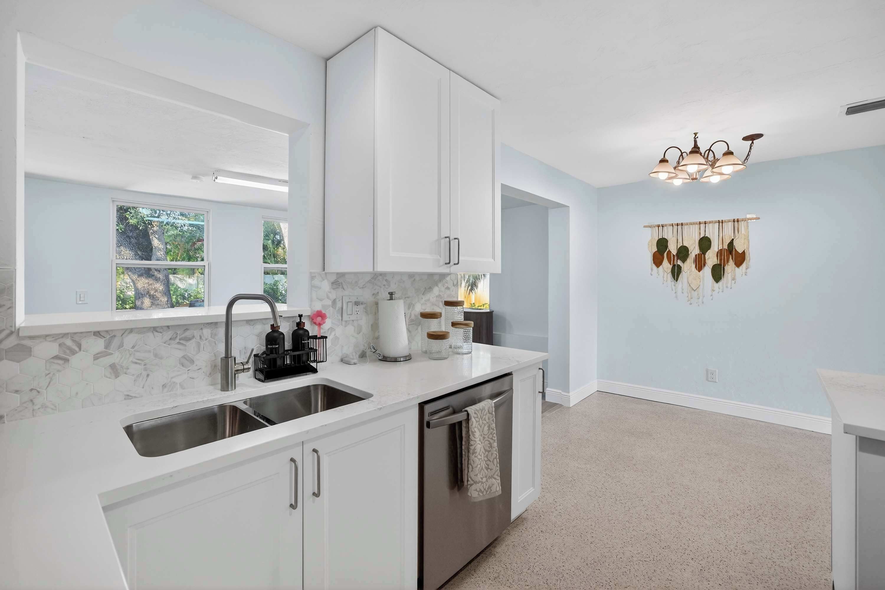 A modern kitchen with white cabinets, a stainless steel sink, and a dishwasher. There is a window providing natural light and a decorative wall hanging.