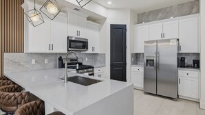 Une cuisine moderne avec des meubles blancs, un îlot central doté d’un évier, des appareils en acier inoxydable et des luminaires suspendus.