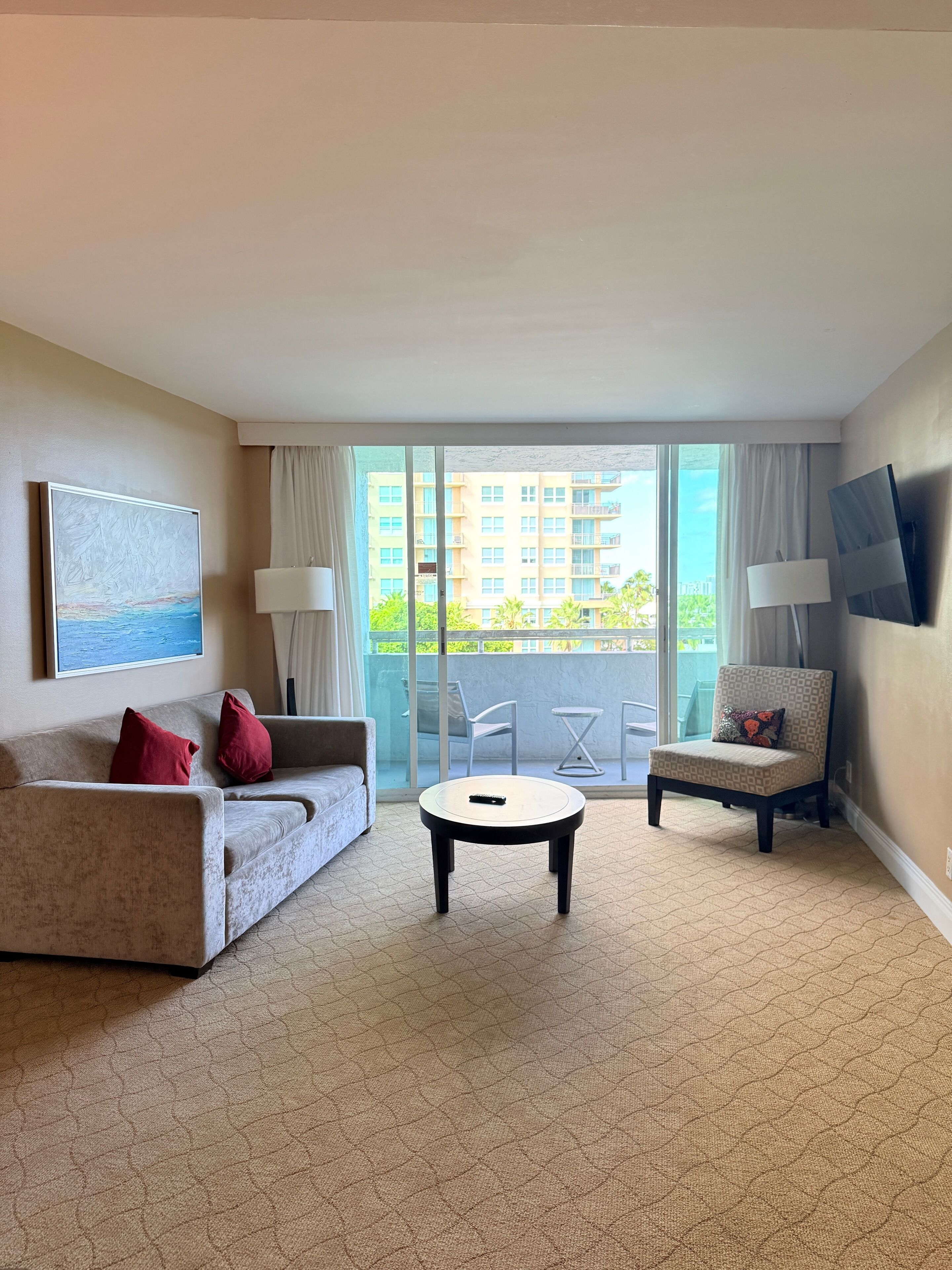 Habitación de hotel con sofá, una silla, una mesita de centro y un ventanal con vistas a edificios y zonas verdes.