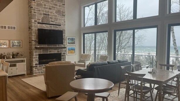 A spacious living room with a stone fireplace, a large dining table, and a view of the outdoors through large windows. - Lanes End: Spacious Lakefront Retreat in Scenic Grand Marais (Grand Marais)