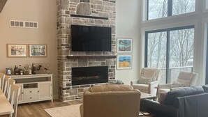 A living room with a stone fireplace, a flat-screen TV, a dining area with a wooden table and chairs, and a large window with a view of trees. - Lanes End: Spacious Lakefront Retreat in Scenic Grand Marais (Grand Marais)