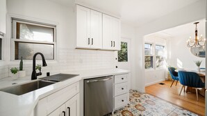A modern kitchen with white cabinets, a stainless steel sink, and a dishwasher. There is a window above the sink and a dining area with a blue chair and a chandelier. - Little Italy Delight! 3 bedroom, backyard, parking (San Diego)