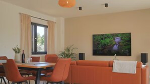 A modern living room with a large orange sofa, a dining table with chairs, a TV mounted on the wall, and a window with curtains. - Villa in Paradise With Heated Pool (Sveta Nedelja)