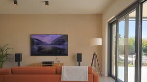 A modern living room with a large window, a sofa, a flat-screen TV, and a floor lamp. - Villa in Paradise With Heated Pool (Sveta Nedelja)