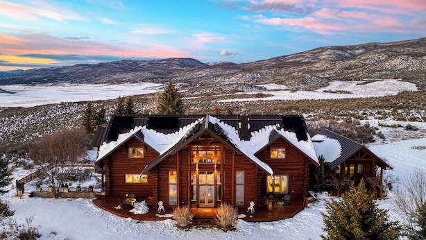 A log cabin with a snow-covered roof, a porch with outdoor seating, and a view of a snowy landscape with mountains in the background. - Sleeps 20, Secluded 7bdr Retreat on 71 Priv Acres Elkstone Lodge by Avantstay (Glenwood Springs)