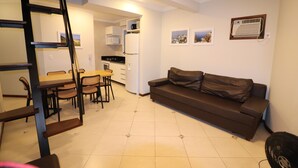 Una sala de estar con un sofá marrón, una mesa de comedor con sillas, una zona de cocina con refrigerador y microondas, y una escalera.