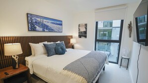 A modern hotel room with a large bed, a nightstand with a lamp, a small table, and a view of the city through a large window. - Mackenzie Mountain Hop (Revelstoke)