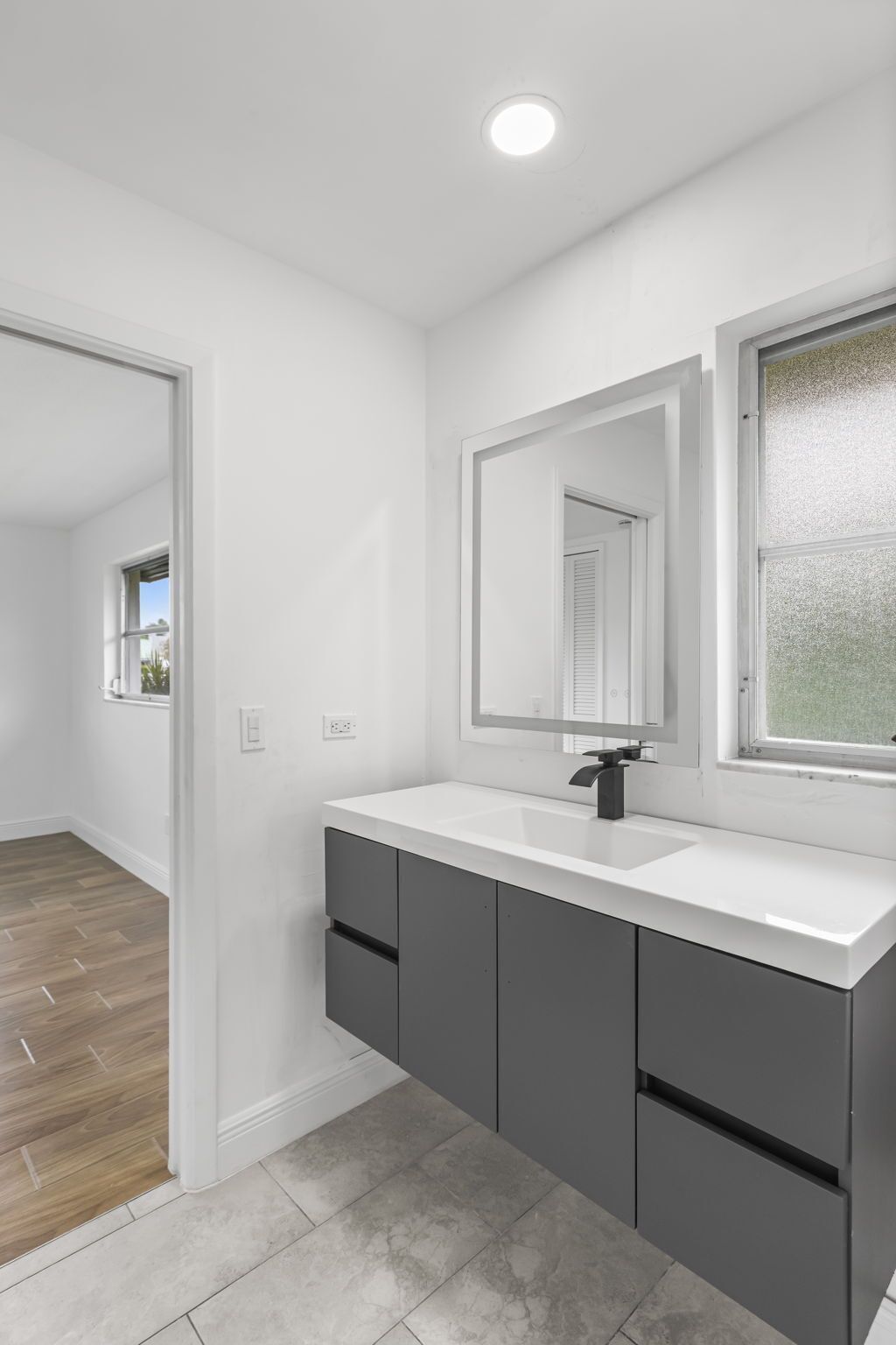 Un baño moderno con tocador blanco, armarios oscuros, un espejo grande y una ventana con vidrio esmerilado.