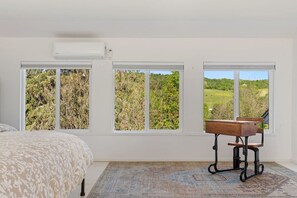 Ein Schlafzimmer mit einem Bett, einem Schreibtisch mit Stuhl und drei Fenstern mit Blick ins Grüne.