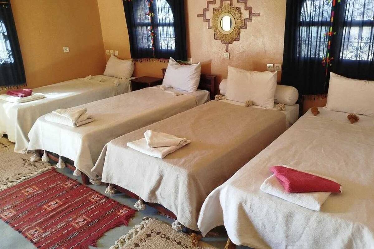 A hotel room with four single beds, each with a white bedspread and pillows, a red patterned rug, and a decorative wall mirror.