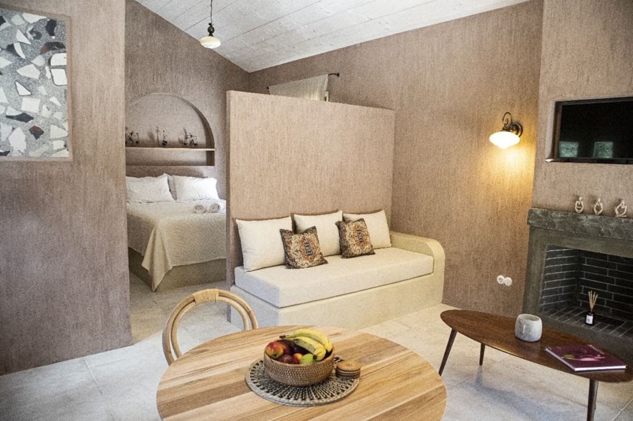 A cozy room with a bed, a sofa, a dining table with fruit, a fireplace, and a wall-mounted TV.