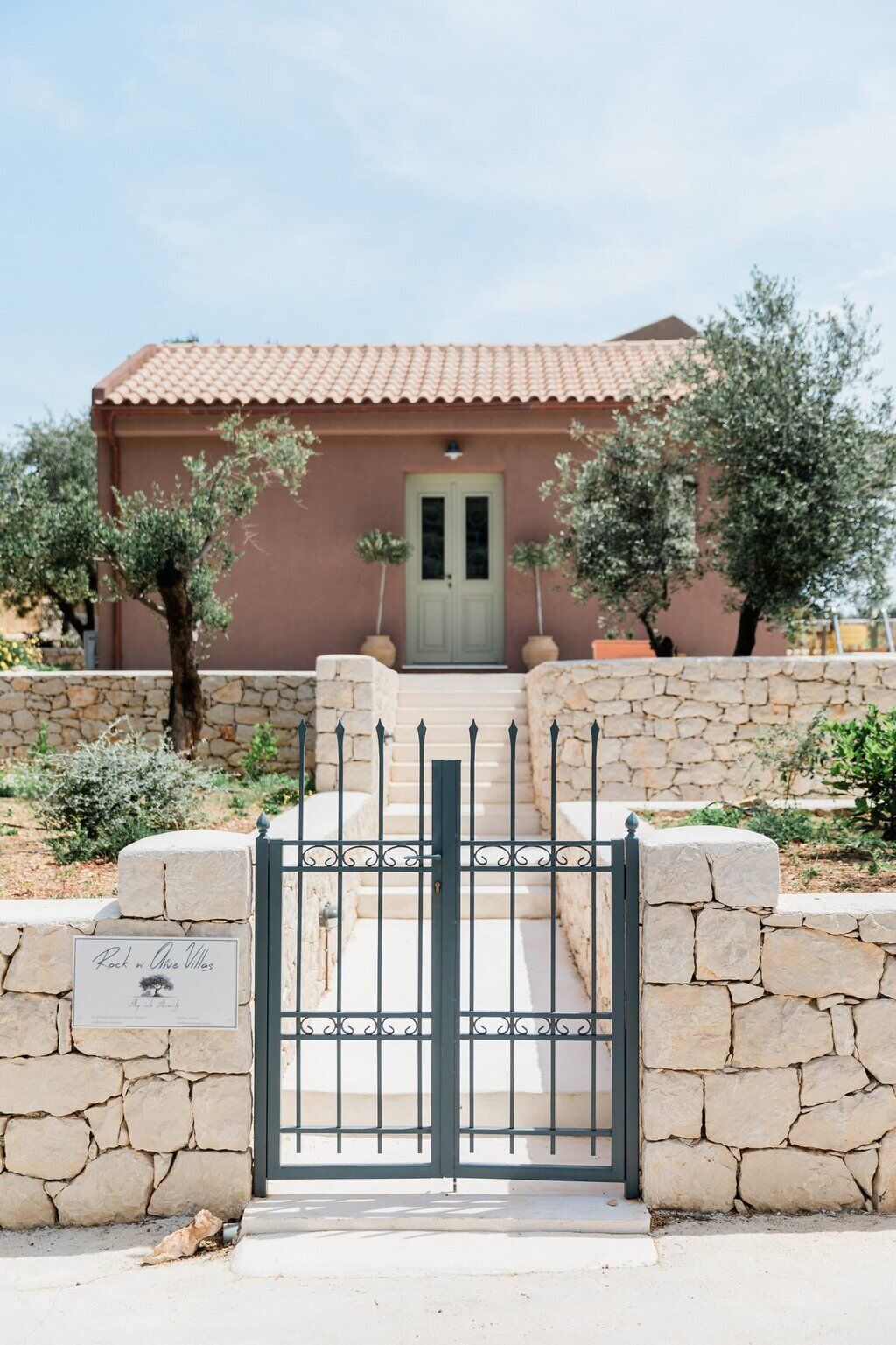 A stone-walled building with a tiled roof, a green door, and a gate with a sign reading "Rock in Olive Valley."