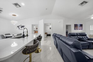 A modern living room with a large sofa, a dining area with a long table, and a kitchen area with a bar. - Cape Coral Oasis |Pool | Fire Pit | GameRoom (Cape Coral)