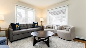 A living room with a grey sofa, a round coffee table, and a grey armchair. There are two windows with blinds, a lamp, and a patterned rug. - Emerald Clover<>3BR, 1Bath House- 1 mile from ND (South Bend)