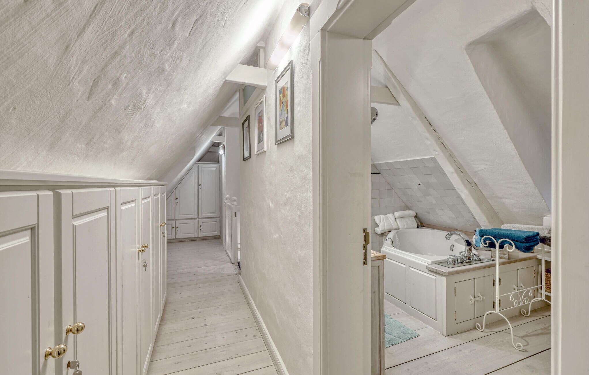 A narrow hallway with white walls and wooden floors leading to a bathroom with a bathtub, towels, and a shower area.