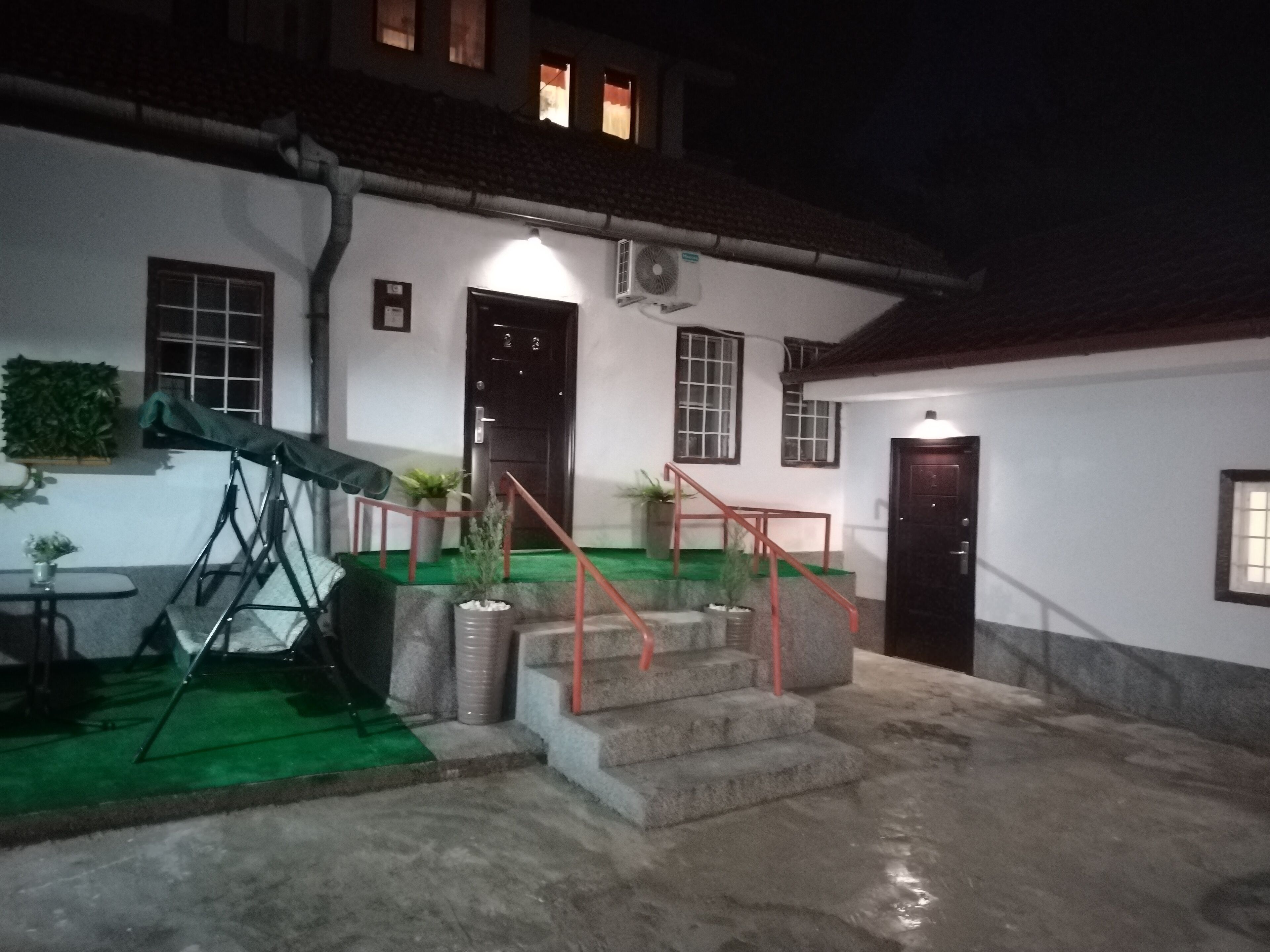 Un edificio con una escalera que lleva a una puerta, un pequeño espacio al aire libre con una mesa y sillas, y un área cubierta con plantas.