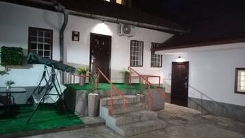 Un edificio con una escalera que lleva a una puerta, un pequeño espacio al aire libre con una mesa y sillas, y un área cubierta con plantas.