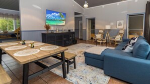 A spacious living area with a wooden dining table, blue sofa, and a television displaying a beach scene.