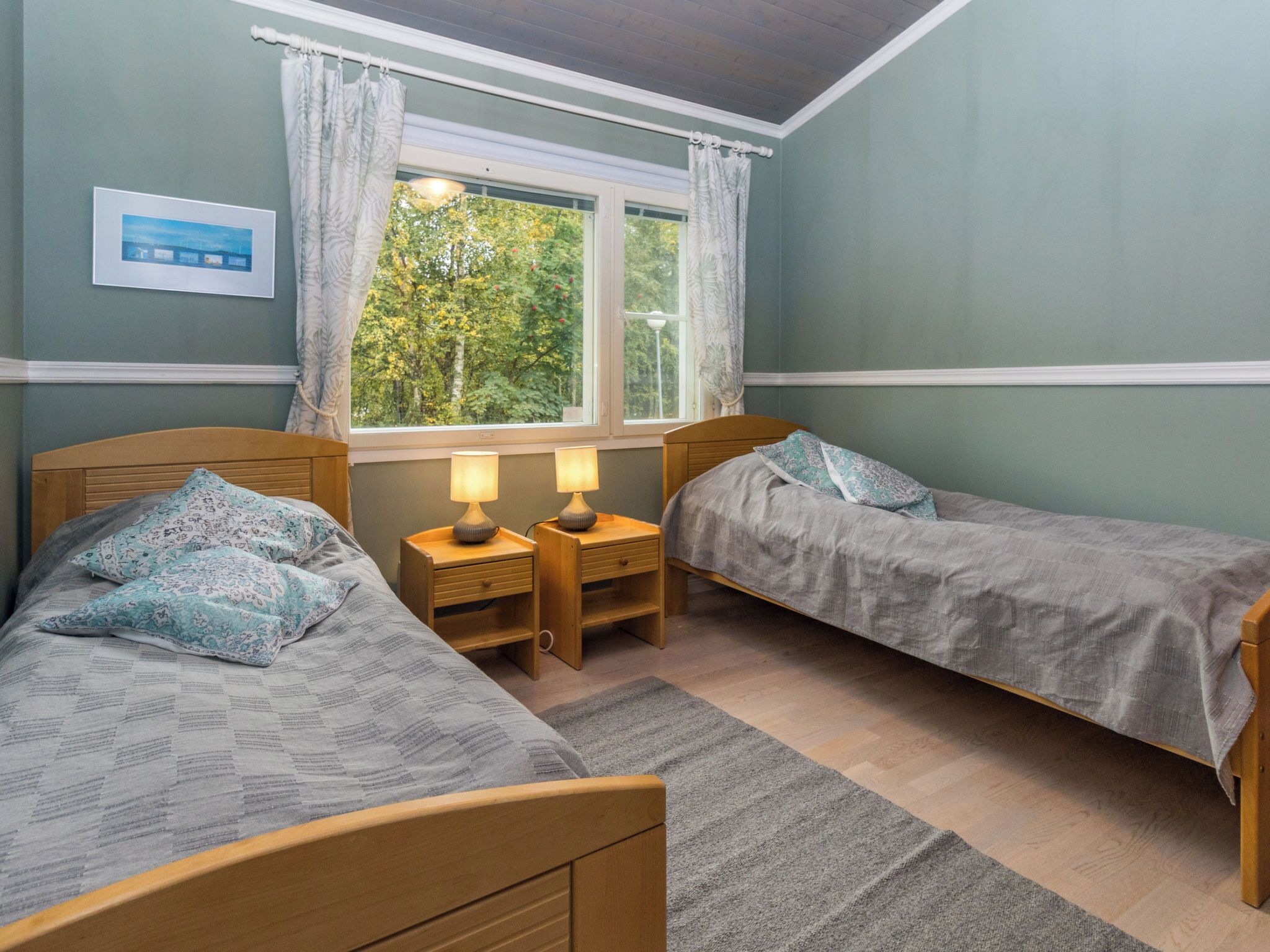 Un dormitorio con dos camas individuales, una ventana con cortinas, una mesita de noche con una lámpara y un cuadro enmarcado en la pared.