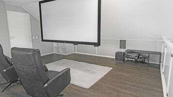 A home theater room with a large flat-screen TV, two gray recliner chairs, a white rug, and a sound system.