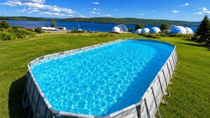 A rectangular swimming pool with clear blue water, surrounded by a grassy area and a view of a lake with mountains in the distance. - Baddeck Domes – Glamping in Nova Scotia (Baddeck)