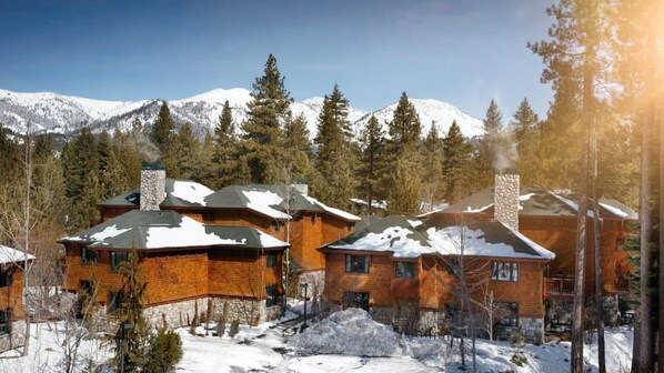 A cluster of log cabins with stone chimneys, surrounded by snow-covered ground and pine trees, with mountains in the background.