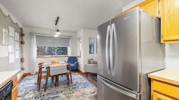 A compact kitchen with a stainless steel refrigerator, wooden cabinets, a dining table with chairs, and a view through a window.