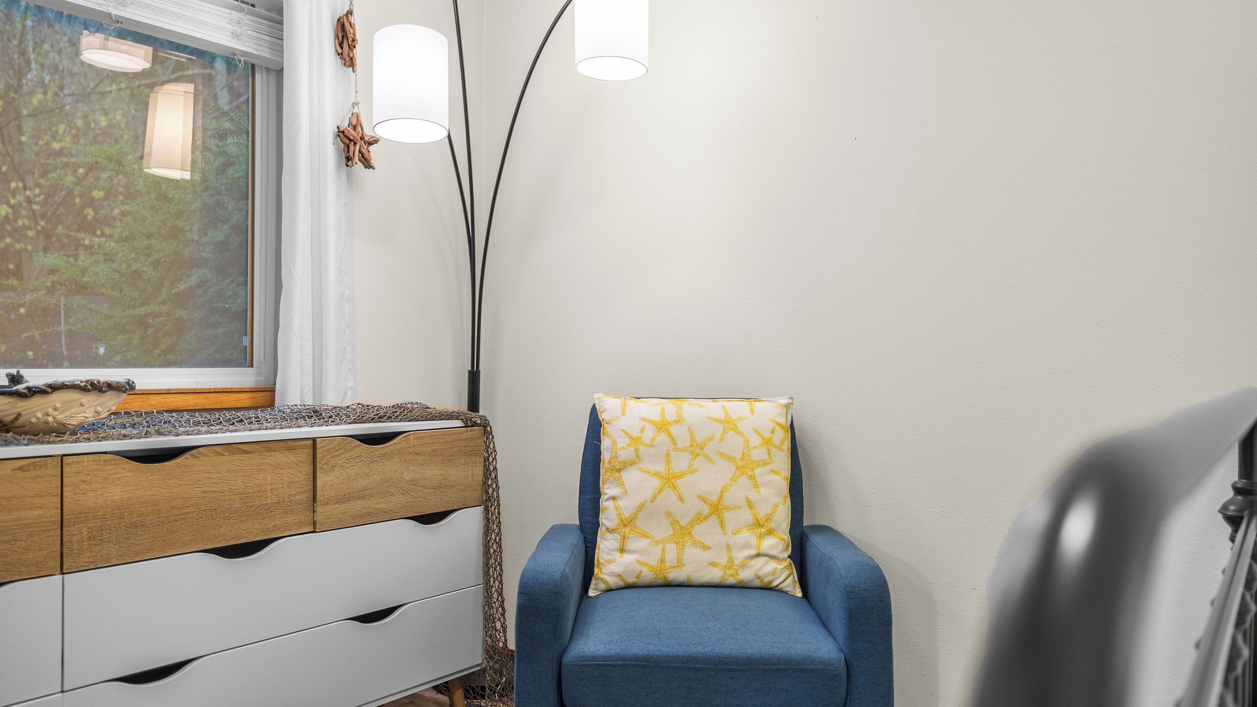 Una habitación con un aparador de madera, una silla azul con cojín estampado, una ventana con vista a follaje y una lámpara fijada a la pared.
