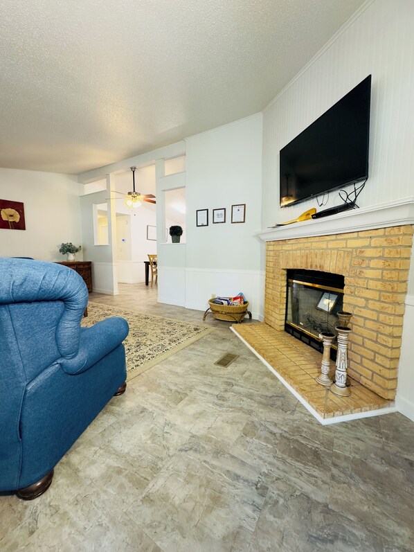 A living room with a blue armchair, a fireplace, a flat-screen TV, and a hallway leading to another room. - Restful, 2b/1b Unit along the Gasconade River (Richland)