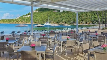An outdoor seating area with tables and chairs, overlooking a beach and the sea.
