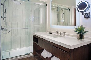 Uma casa de banho moderna com cabine de duche em vidro, lavatório branco com duas torneiras e espelho com luz redonda.