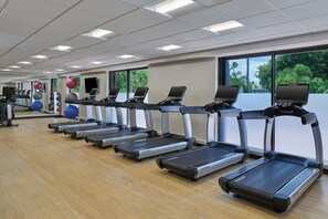 Uma academia com várias esteiras alinhadas, bolas de exercício e uma grande janela com vista para o verde.