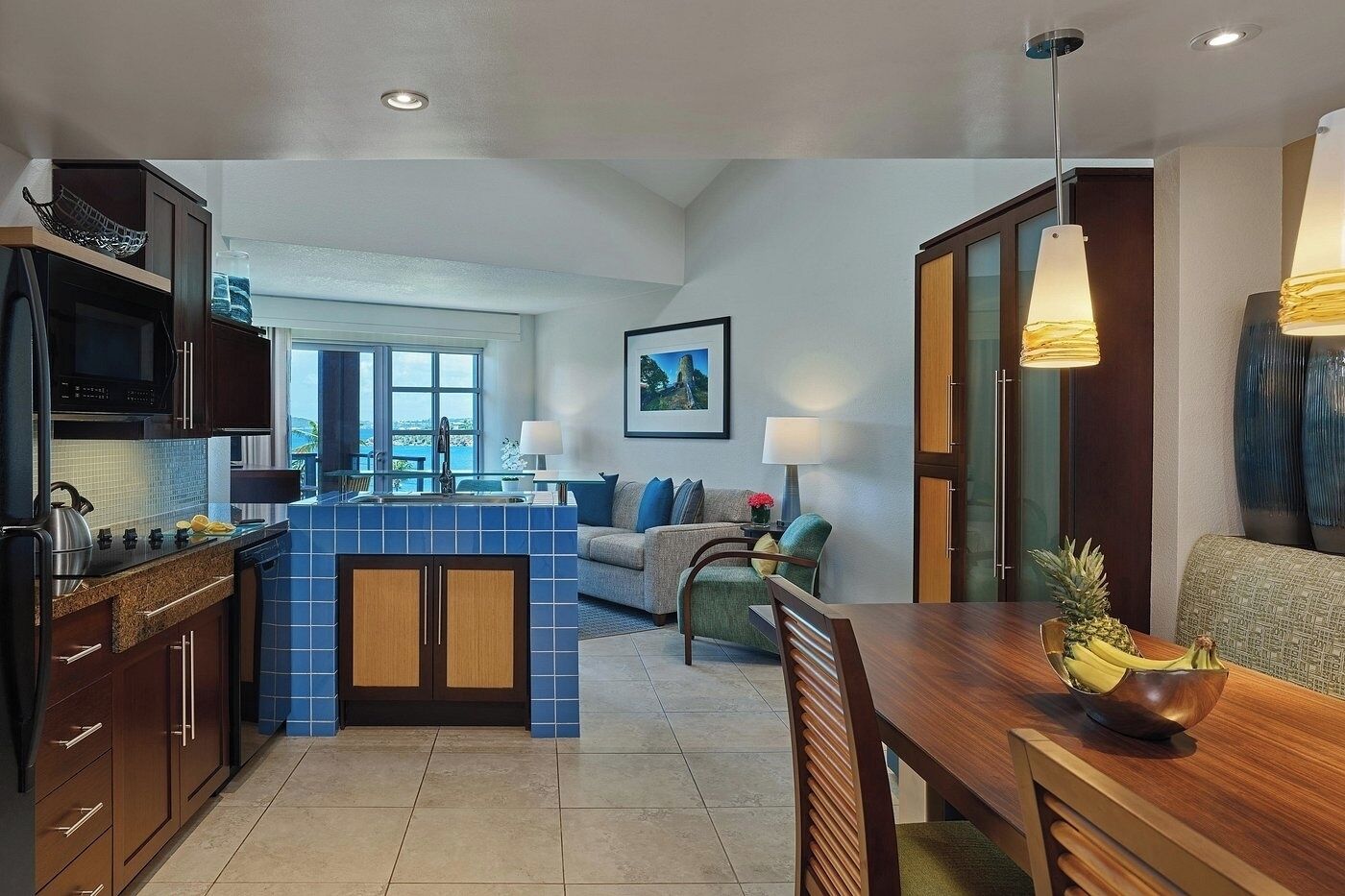 A modern kitchen with blue tiles, a dining area with a wooden table, and a living room with a sofa and a painting on the wall.