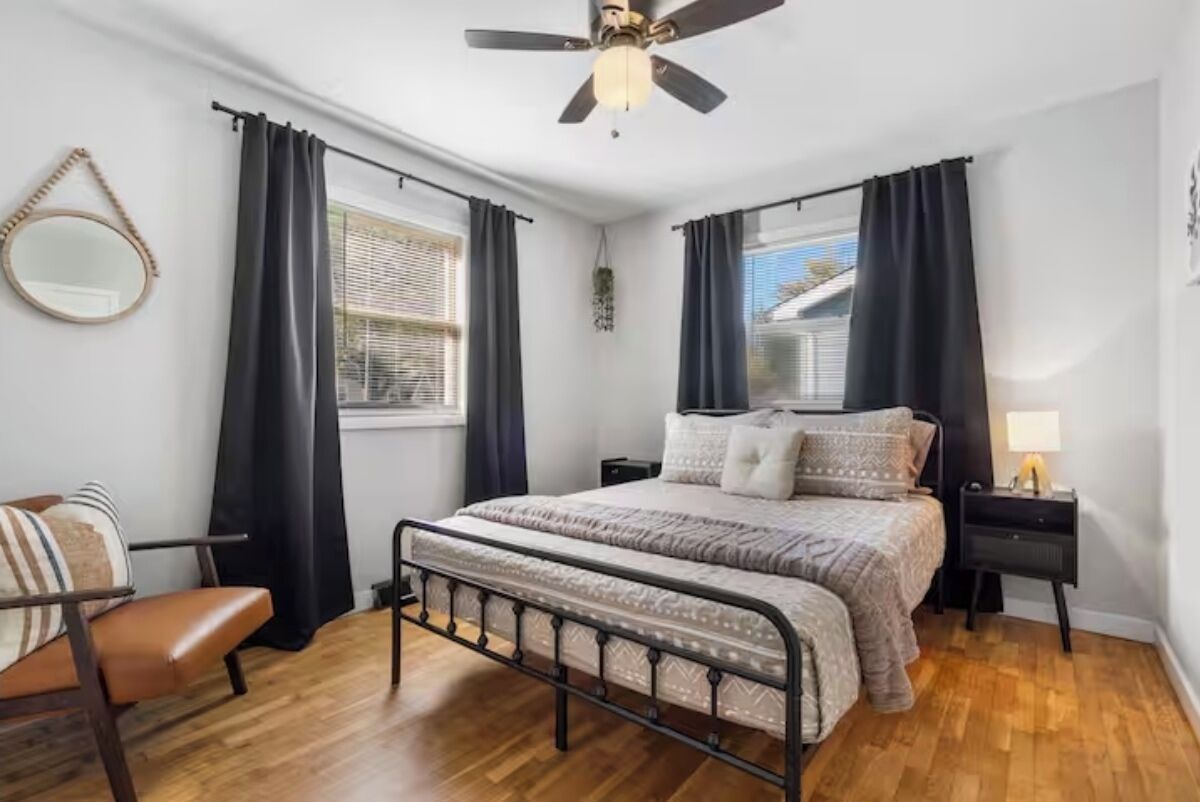A bedroom with a bed, a chair, a bedside table, a ceiling fan, and a window with curtains.