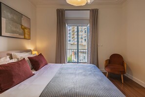 Ein Hotelzimmer mit einem großen Bett, einem Sessel, einem Gemälde und Blick auf ein Gebäude.