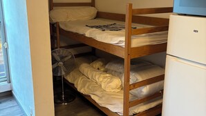 Een stapelbed met een televisie erboven, een ventilator en een koelkast in een kamer met houten vloeren en een raam.
