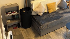 Een kleine kamer met een grijze slaapbank, een klein houten plankje, een zwarte prullenbak en een witte wasmachine.