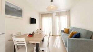 Una sala de estar moderna con un sofá gris, una mesa de comedor de madera, sillas blancas y un televisor montado en la pared.