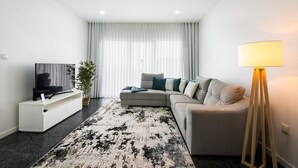 Uma sala de estar moderna com um sofá modular, uma TV de tela plana sobre um suporte branco, um tapete estampado, um abajur de chão e uma janela grande com cortinas finas.