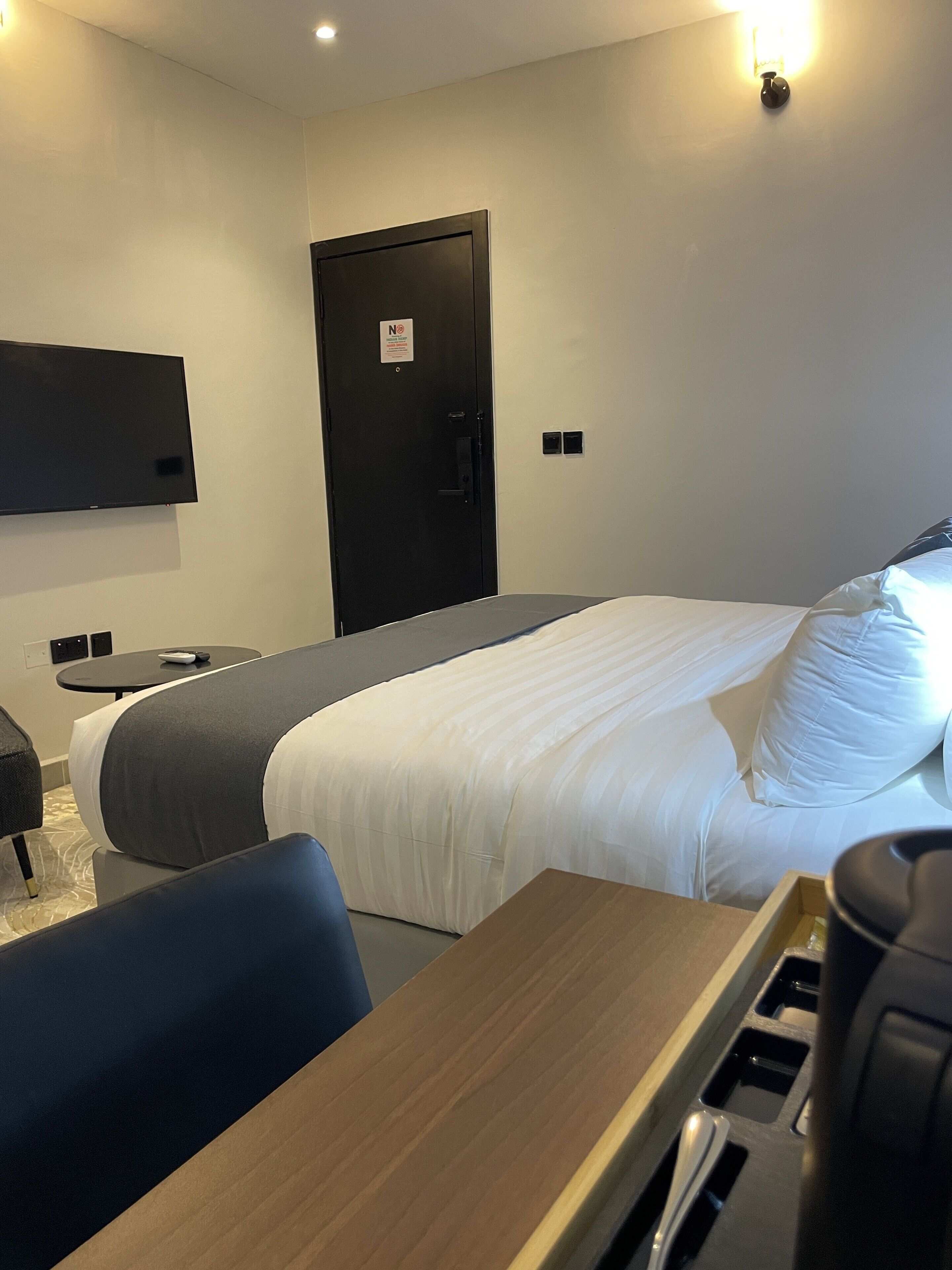 A hotel room with a bed, a desk, a chair, a TV mounted on the wall, and a door with a sign.
