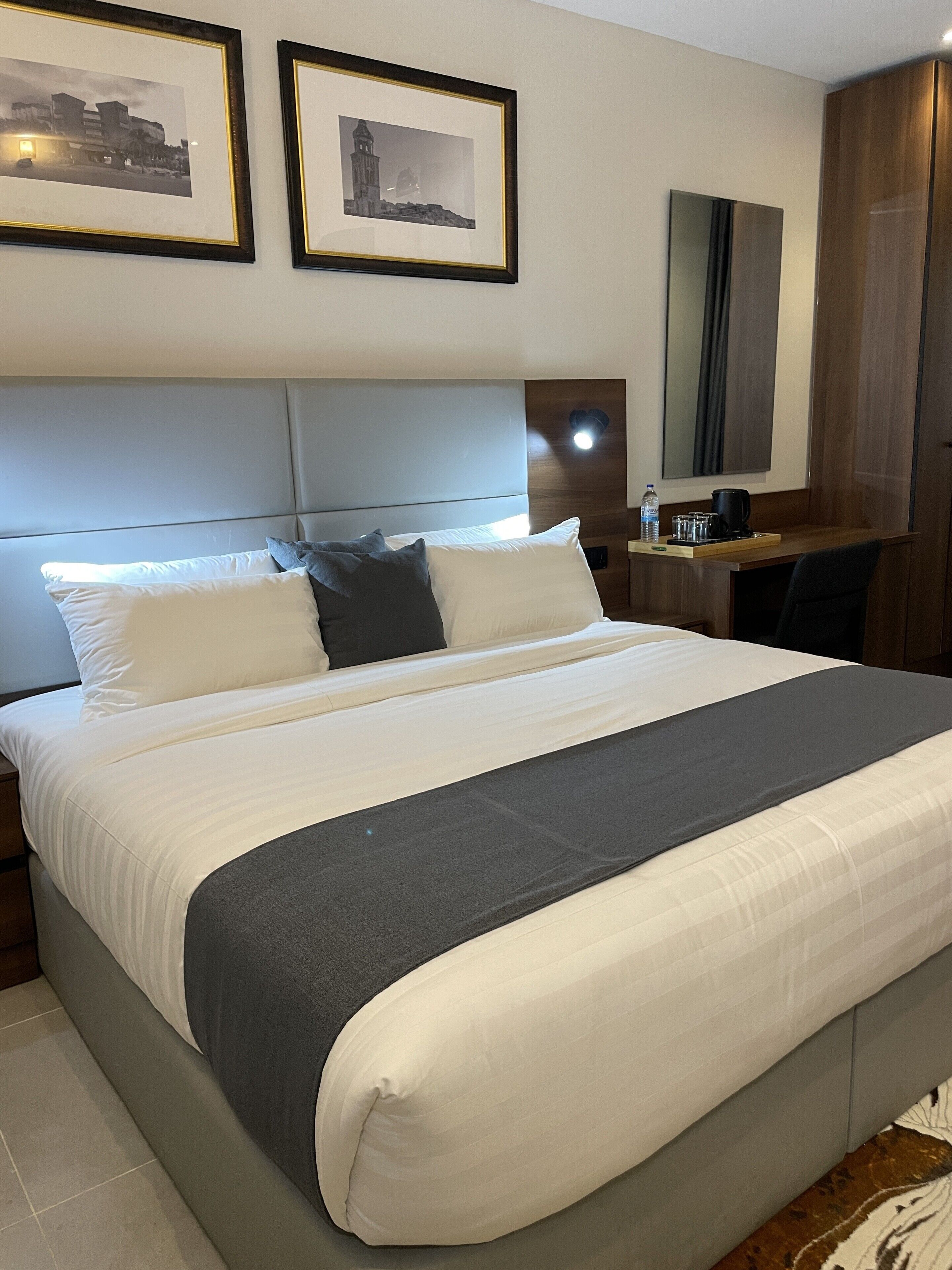 Een net opgemaakt bed met wit en grijs beddengoed, een hoofdbord en een nachtkastje met een waterfles en een koffiezetapparaat.