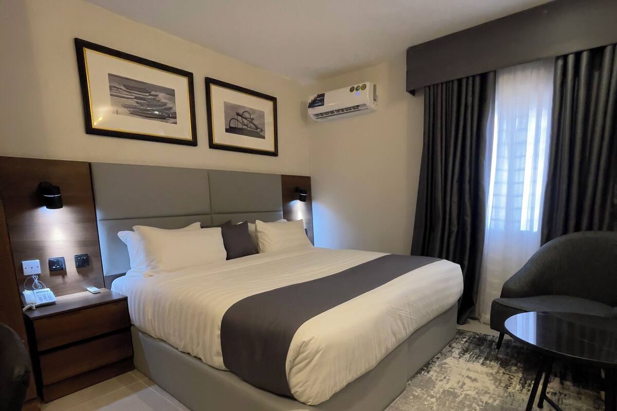 A hotel room with a bed, bedside tables, a desk, a chair, an air conditioning unit, and two framed pictures on the wall.