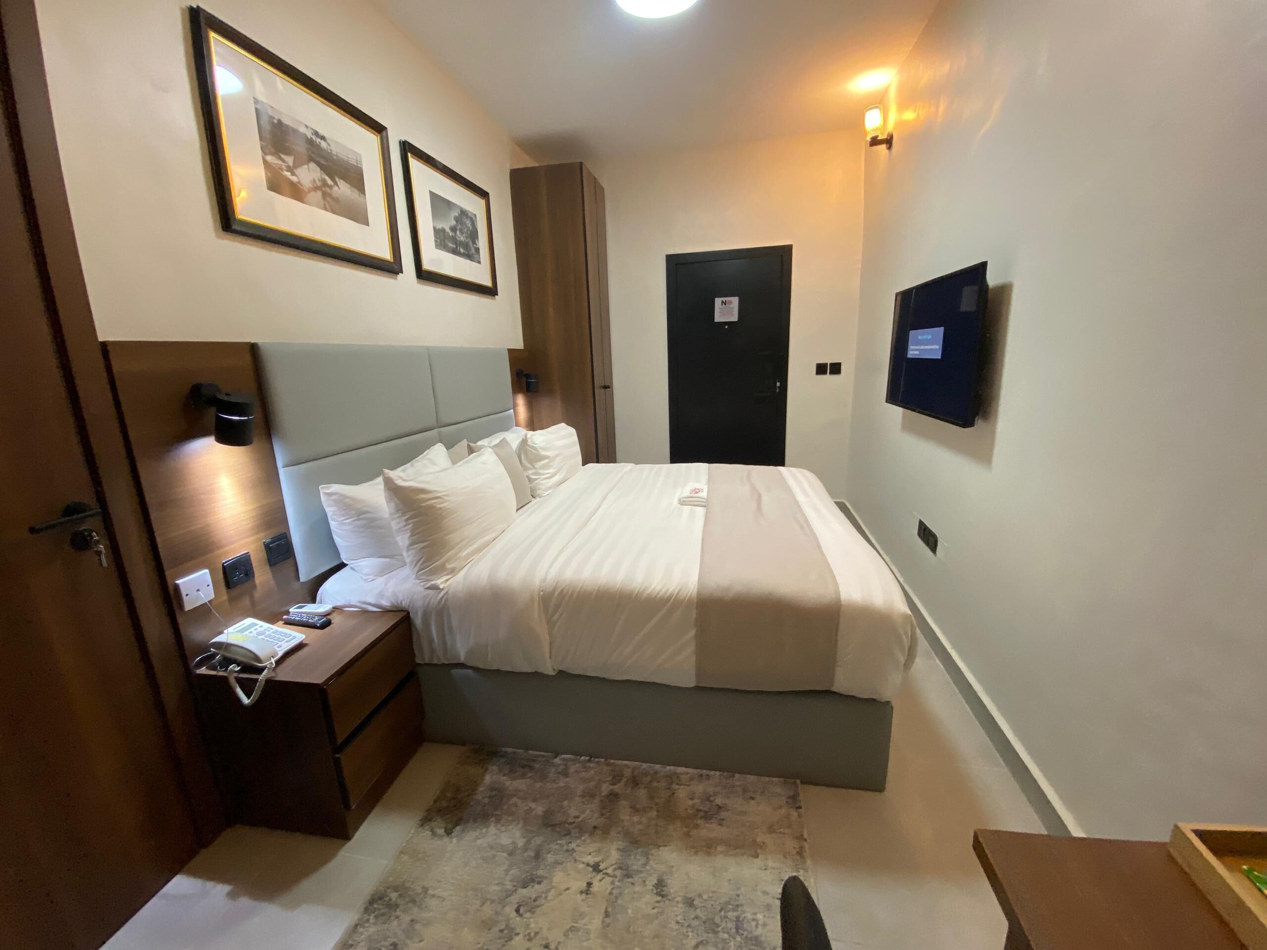 Een hotelkamer met een bed, nachtkastje, lamp, ingelijste foto's en een televisie aan de muur.