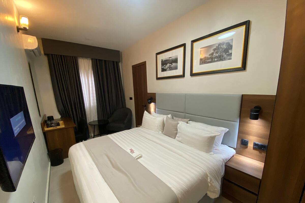 A hotel room with a large bed, a desk, a chair, a TV, and two framed pictures on the wall.